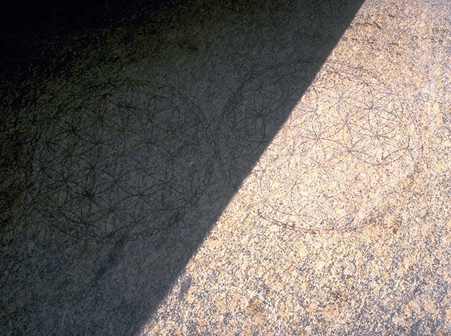 Dessin gravé en pierre montrant une motif de fleur de vie entouré de motifs géométriques, partiellement obscurci par une ombre diagonale.